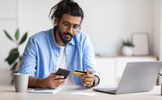 Casual man looking at cell phone and credit card next to laptop and cup of coffee