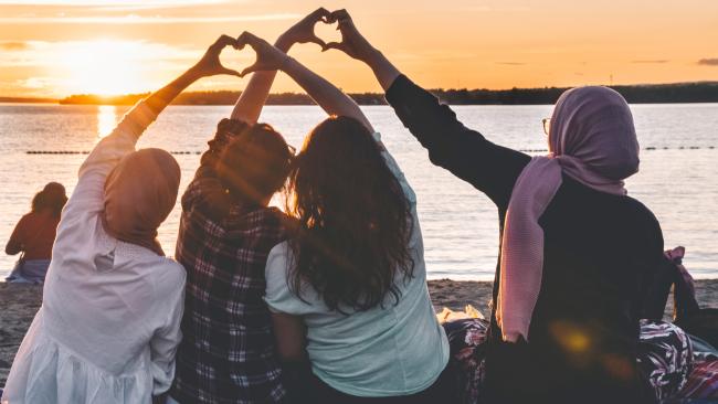 women making heart with hands by lake at sunset